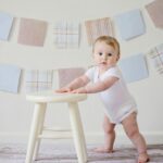 Cute baby standing by a stool indoors with a playful background.